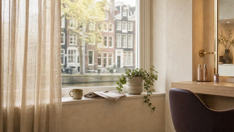 Zicht vanaf een stoel in de salon door het raam op Amsterdamse grachtenpanden, met een mok en een plant op de vensterbank