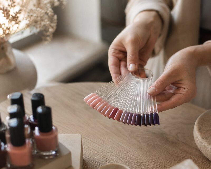 Handen houden een waaier met gellak-kleurstaaltjes boven een lichte salontafel, met nagellakflessen op de achtergrond
